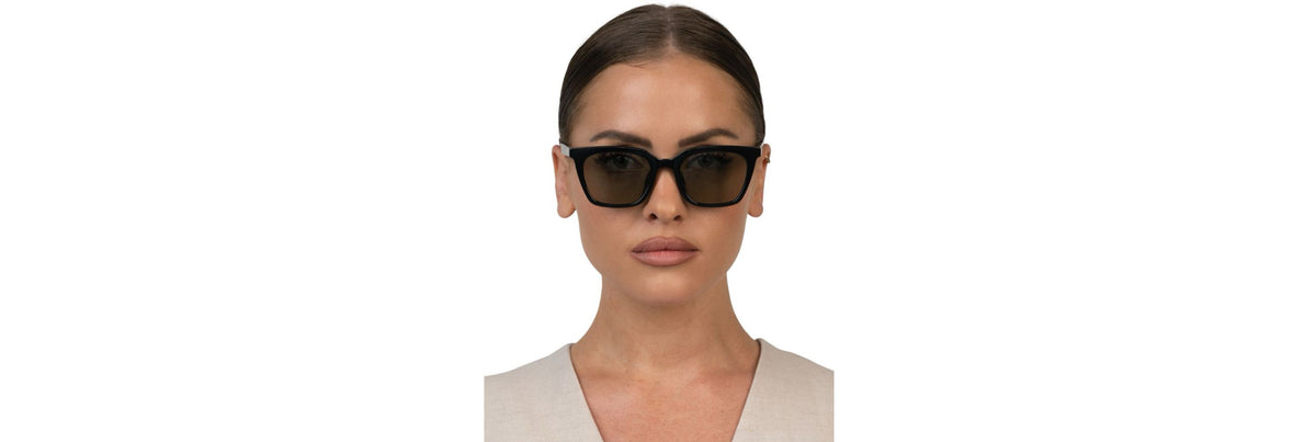 Close-up of UV400 sunglasses held against bright sun, showing a medium-brown fade lens and full eye coverage around the brow and temples.