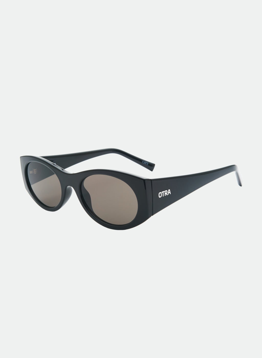 Otra Eyewear - GRACE - Black/Smoke - Angled view of GRACE sunglasses in Black/Smoke featuring a glossy black oval frame, smoke-tinted lenses, and a softly curved silhouette, displayed against a clean light grey background. A timeless everyday style designed for versatile wear with full UV400 protection.