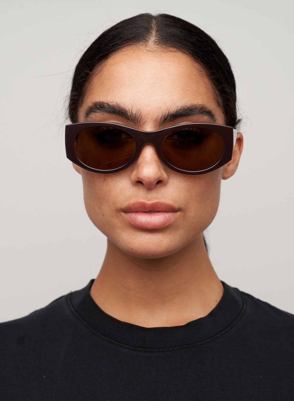 Otra Eyewear - GRACE - Dark Chocolate/Brown - Model wearing of GRACE sunglasses in Dark Chocolate/Brown featuring a glossy dark chocolate oval frame, warm brown lenses, and a softly curved silhouette, displayed against a clean light grey background. A timeless everyday style designed for versatile wear with full UV400 protection.