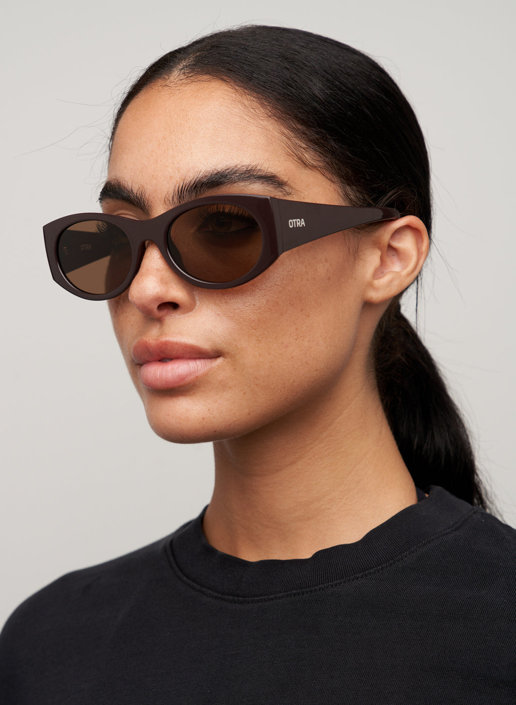 Otra Eyewear - GRACE - Dark Chocolate/Brown - Front view of GRACE sunglasses in Dark Chocolate/Brown featuring a glossy dark chocolate oval frame, warm brown lenses, and a softly curved silhouette, displayed against a clean light grey background. A timeless everyday style designed for versatile wear with full UV400 protection.