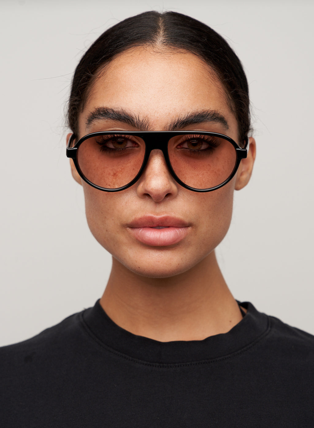 Otra Eyewear - IGGY - Black/Light Brown Fade - Model wearing a IGGY sunglasses in Black/Light Brown Fade featuring a high-gloss black aviator frame, classic double bridge, and light brown gradient lenses, displayed against a clean grey background. A timeless everyday design with modern styling and full UV protection 