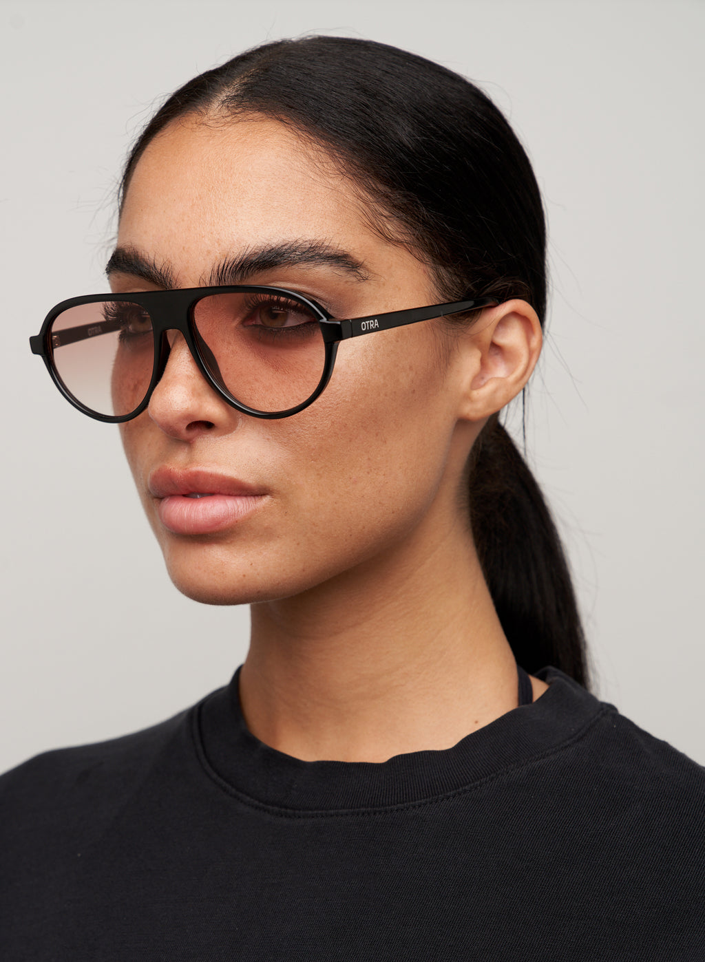 Otra Eyewear - IGGY - Black/Light Brown Fade - Model wearing of IGGY sunglasses in Black/Light Brown Fade featuring a high-gloss black aviator frame, classic double bridge, and light brown gradient lenses, displayed against a clean grey background. A timeless everyday design with modern styling and full UV protection 