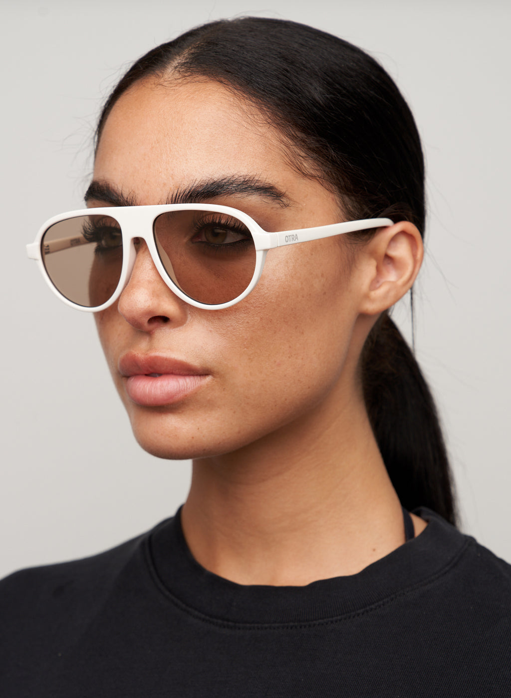 Otra Eyewear - IGGY - Vanilla/Brown - Model wearing IGGY sunglasses in Vanilla/Brown featuring a high-gloss vanilla cream aviator frame, classic double bridge, and light brown lenses. Front-facing editorial portrait showcasing the timeless everyday silhouette with a clean, modern aesthetic in grey background.