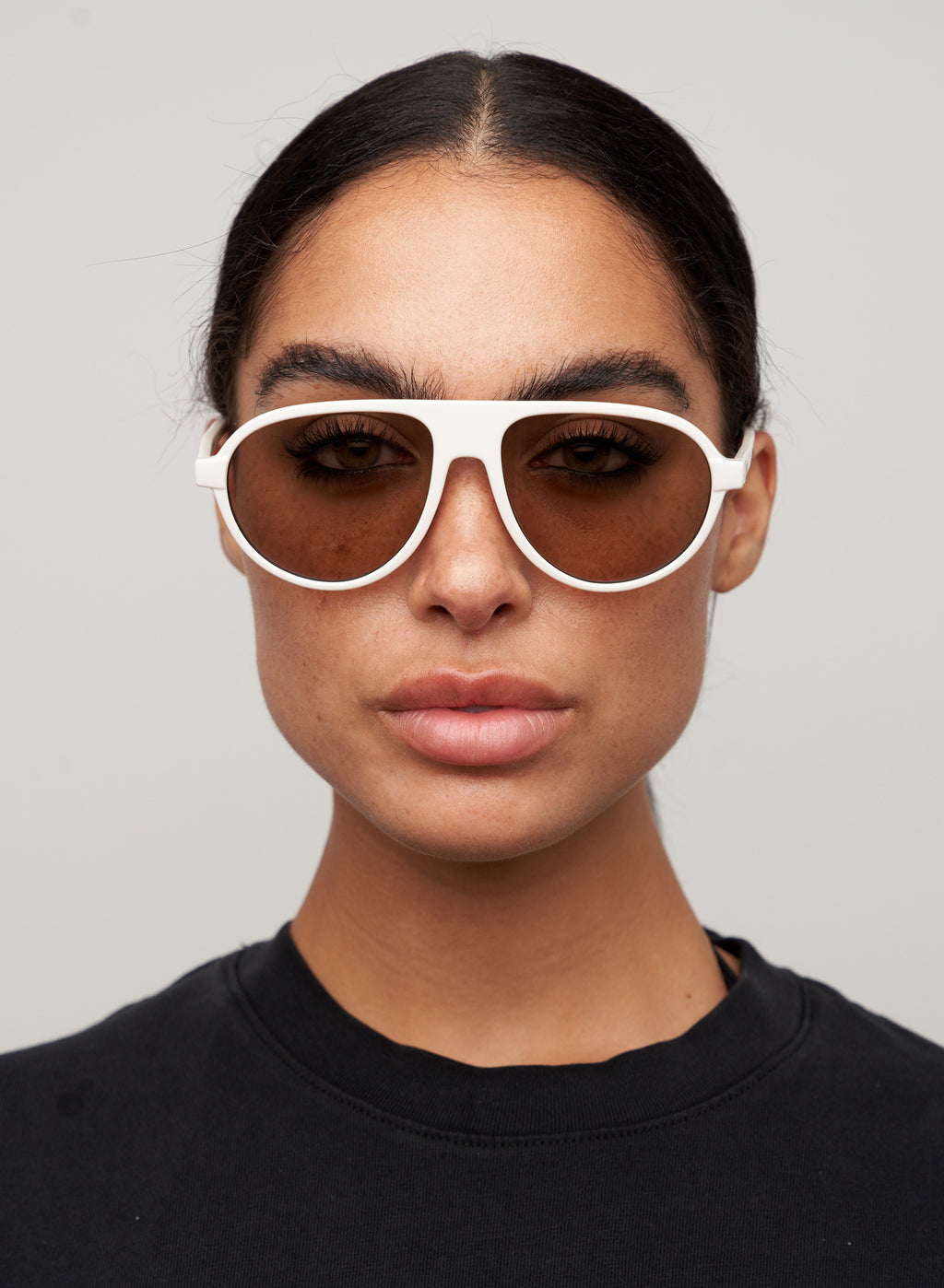 Otra Eyewear - IGGY - Vanilla/Brown - Model wearing IGGY sunglasses in Vanilla/Brown featuring a high-gloss vanilla cream aviator frame, classic double bridge, and light brown lenses. Front-facing editorial portrait showcasing the timeless everyday silhouette with a clean, modern aesthetic in grey background.