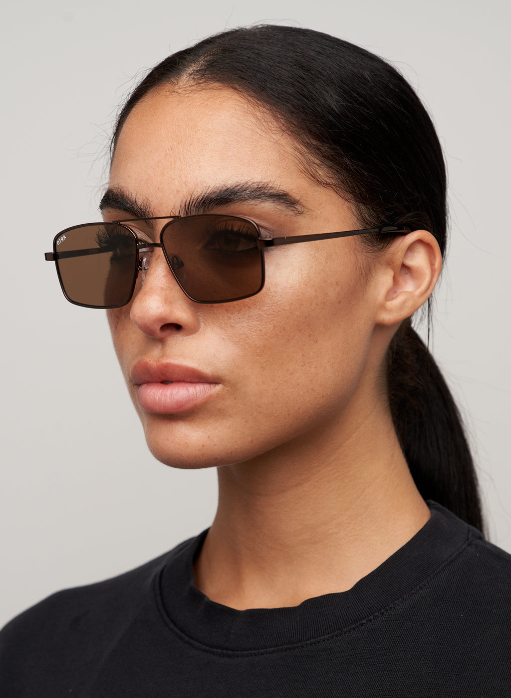 Otra Eyewear - NASH - Chocolate/Brown - Model wearing a NASH sunglasses in Chocolate/Brown featuring a sleek chocolate metal square aviator frame, warm brown lenses, and a refined double-bridge design, displayed against a clean light grey background. A modern aviator style with lightweight construction and timeless everyday appeal.