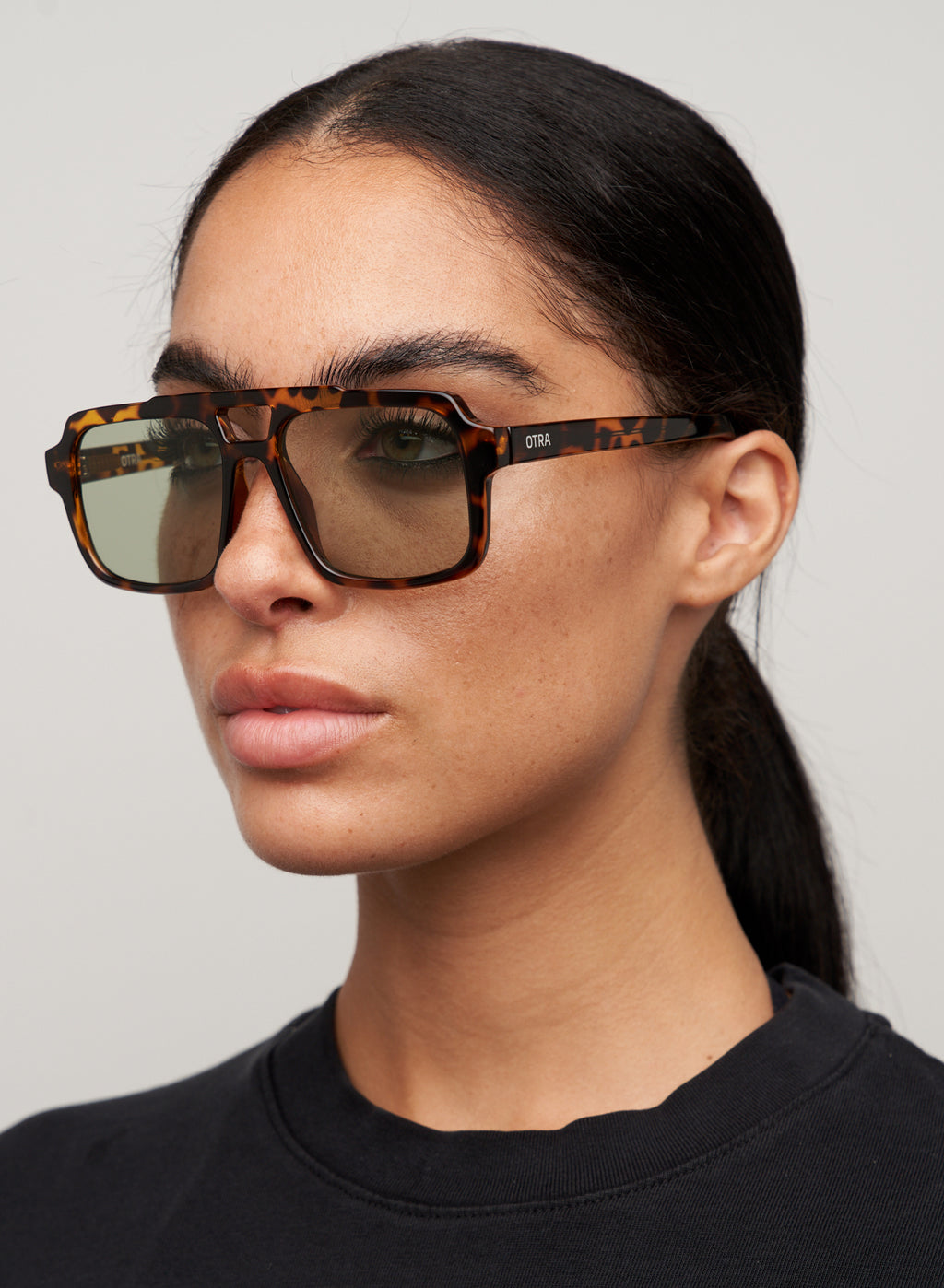 Otra Eyewear - TAME - Tort/Light Green - Model wearing a TAME sunglasses in Tort/Light Green featuring a lightweight tortoiseshell square aviator frame, light green lenses, and a structured angular silhouette. A modern interpretation of the classic aviator designed for effortless everyday style in grey background