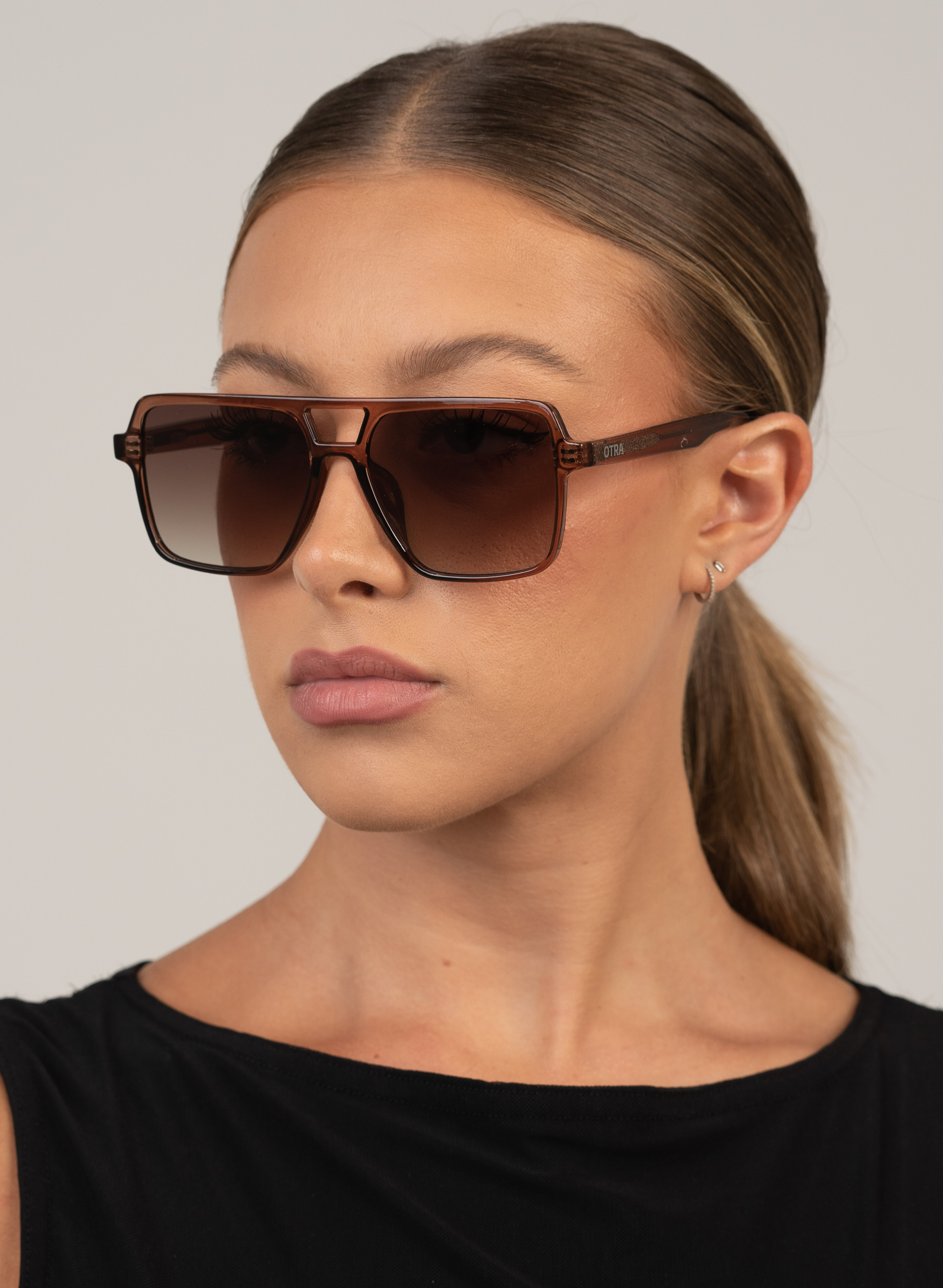 Otra Eyewear- Zoya-Transparent Maple- Three-quarter side portrait view of a model wearing the Zoya oversized square large aviator sunglasses in a warm transparent maple color with brown gradient lenses against a light background.  effortless edge and sun protection to city strolls, road trips, and everything in between.  

